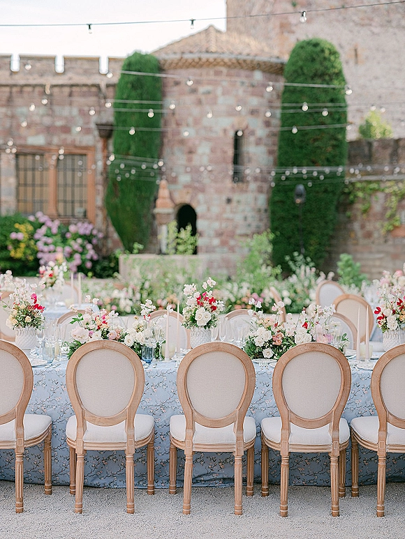 Reception tablescape with a blue floral tablecloth, colorful centerpieces, taper candles, and glassware under string lights in a stone courtyard garden