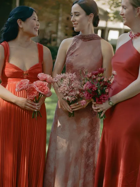 Bridesmaid portrait of women holding bouquets, laughing together in mismatched dresses on a sunlit garden lawn with trees and a building behind