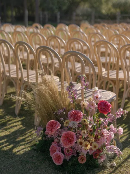 Ceremony aisle decor with outdoor ceremony seating, wood chairs flanking a pink rose and wildflower ground arrangement on a sunny lawn