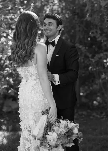 First look moment as bride in lace spaghetti-strap gown touches groom’s face; he wears black tux with bow tie in dappled garden light