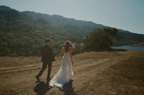 Couple portrait of bride and groom walking away holding hands, her lace gown train trailing, with mountains and lake under clear sky
