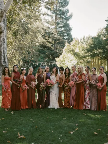Bridesmaid group photo with bride, bridesmaids in rust dresses holding pink and coral bouquets on a garden lawn under string lights