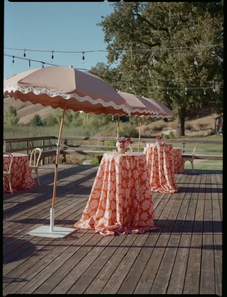 Cocktail hour decor with outdoor cocktail tables draped in floral linens, bud vases, bistro chairs, and string lights on a deck with hills
