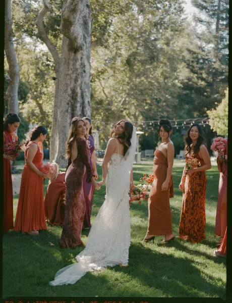 Bride with bridesmaids in rust dresses holding bright bouquets, the bride in veil and lace gown on a garden lawn with string lights