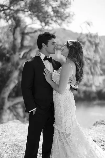 Couple portrait of bride in lace wedding dress embracing groom in tuxedo by rocky lakeshore with cliffs, trees, and sky behind