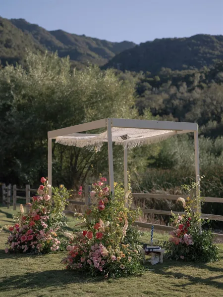 Wedding ceremony arch with a white square frame and fringe canopy, colorful florals and greenery, set on a lawn with mountain views