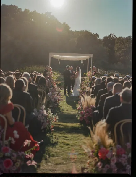 Ceremony moment at an outdoor wedding ceremony with bride and groom under a draped arch, pink florals and pampas, sunlit hillside aisle