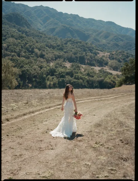 Bridal portrait of a bride walking away and looking back, holding a bouquet in a strapless lace dress on a dirt road with mountains behind