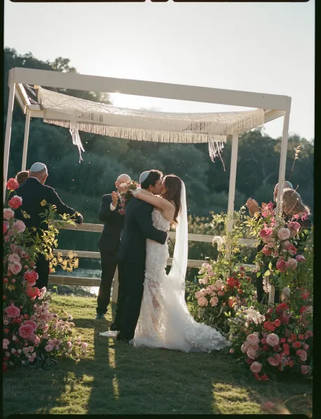 Ceremony kiss as bride in lace dress and long veil kisses groom under a macrame floral arch on a sunlit hillside lawn
