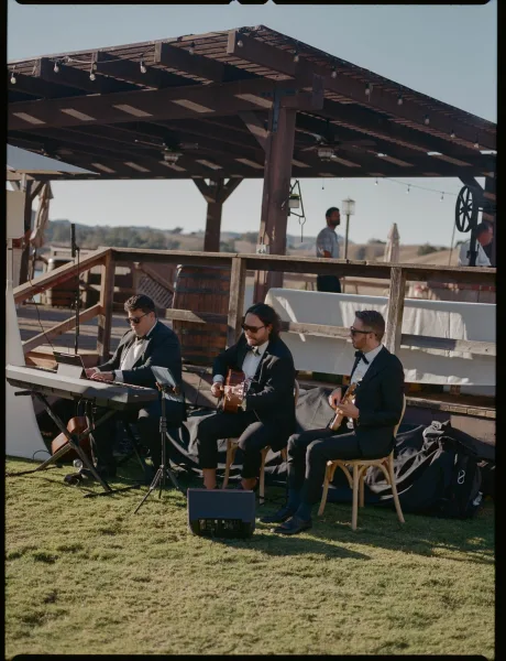Wedding ceremony musicians in black suits and sunglasses perform live wedding ceremony music on a patio deck under pergola string lights