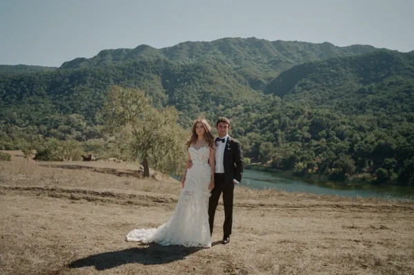 Couple portrait of bride in a strapless lace gown with train and groom in black tuxedo, posing by a mountain lake hillside backdrop