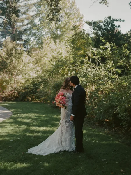 Wedding kiss portrait of bride and groom kissing on a sunlit garden lawn, her colorful bouquet and lace train beside his black suit boutonniere