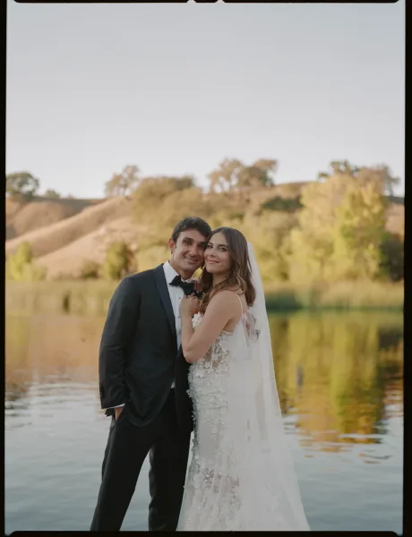 Couple portrait of bride in strapless lace wedding dress and veil embracing groom in black tuxedo by a calm lake with hills and trees