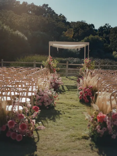 Ceremony aisle decor with pink roses and garden flowers lining a lawn aisle, leading to a white draped arch beside a wooden fence