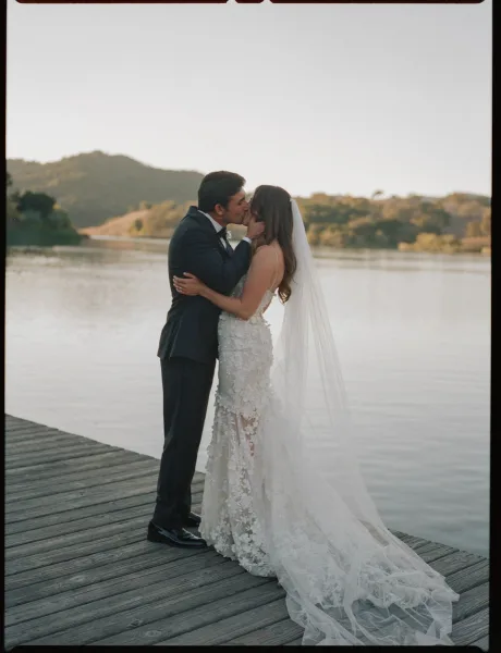 Wedding kiss as the bride and groom kissing on a wooden dock, her lace gown and long veil flowing with a mountain lake behind them