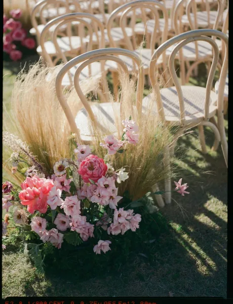 Ceremony seating with outdoor wedding chairs, bentwood rows framing pink floral and ornamental grass clusters on a sunlit garden lawn aisle