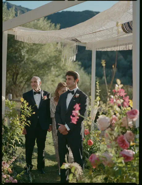 Groom portrait in a black tuxedo with bow tie and boutonniere, standing beneath a fringe canopy in a garden with mountains behind
