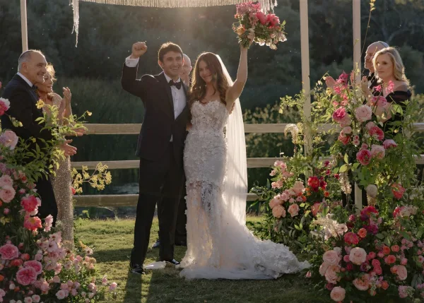Ceremony recessional as newlyweds walk down the aisle, bride lifting her bouquet and groom fist-pumping beneath a fringe canopy on a lawn