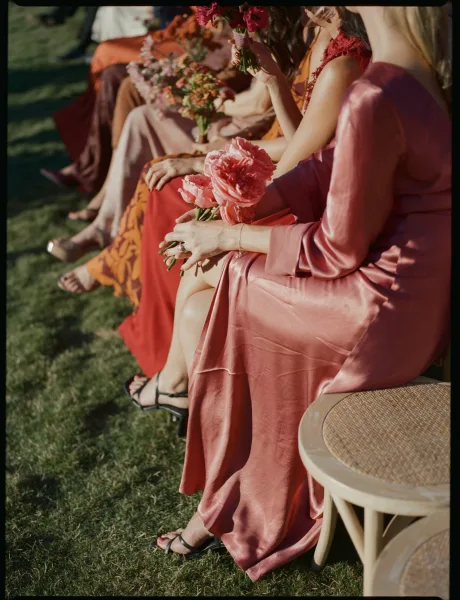 Bridesmaid dresses in dusty rose satin with floral bouquets and strappy heels, seated by wicker chairs on a grass lawn outdoors