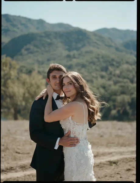 Couple portrait of bride and groom embrace, her lace spaghetti-strap gown against his tuxedo and bow tie in a mountain meadow