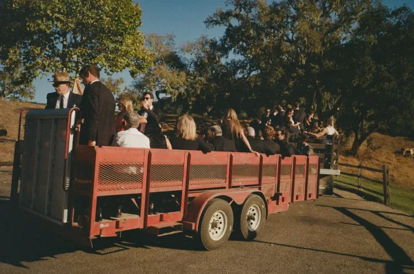 Wedding guest transportation with a tractor pulling an open trailer of guests in suits down a country road with hills, fence, and blue sky