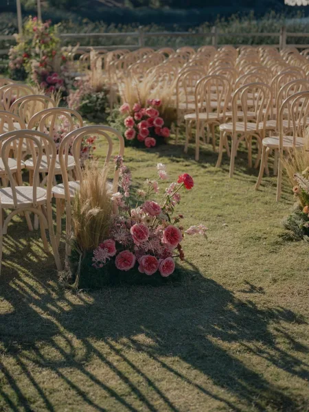 Ceremony aisle decor with outdoor ceremony aisle flowers in low pink rose and wildflower arrangements along wood chairs on a sunlit lawn