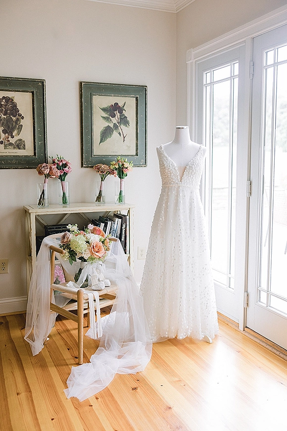 Wedding dress on a mannequin with lace details, bridal veil and bouquet accents in a sunlit room by French doors