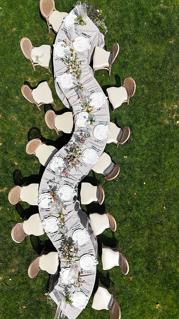 Reception tablescape with an outdoor wedding dinner table in a long serpentine curve, floral garland, bud vases, candles on green lawn grass