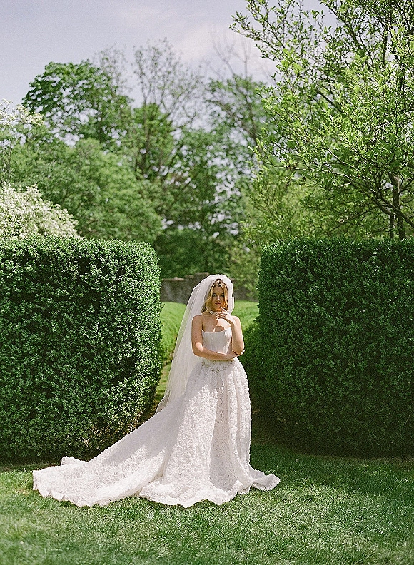 Bridal portrait of a bride holding her cathedral veil, wearing a strapless corset lace gown and necklace before garden hedges and trees