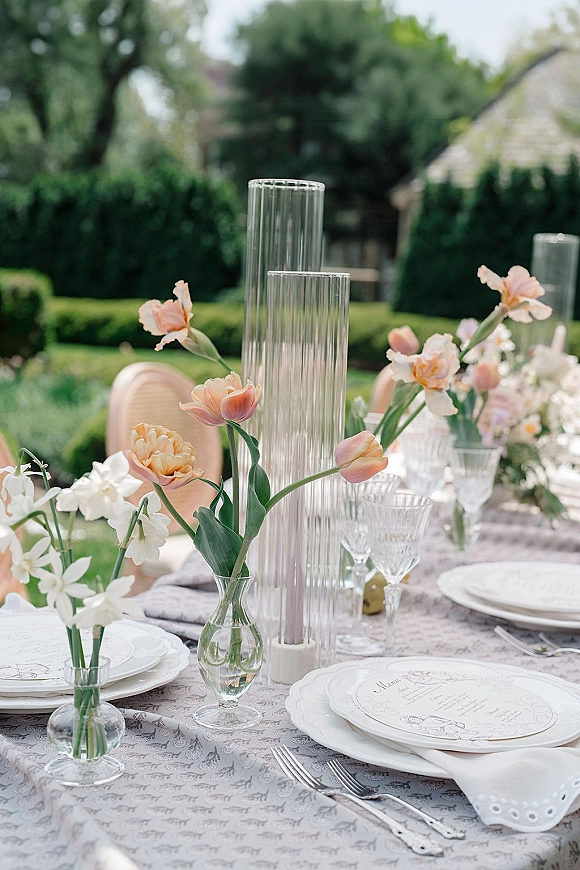 Wedding tablescape with floral centerpieces and bud vases on a patterned tablecloth, outdoor reception table set on a garden patio with rattan chairs