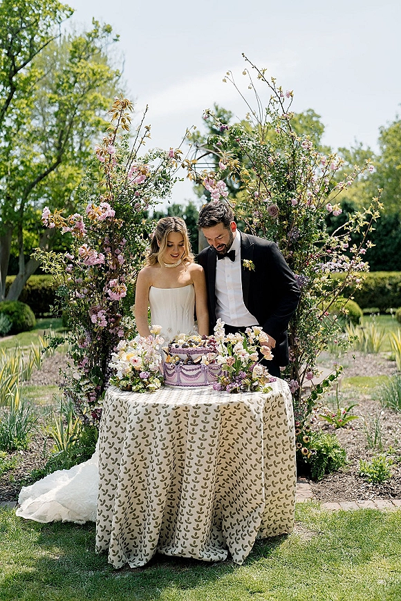 Wedding cake cutting as bride in strapless dress and groom in tux slice a purple buttercream cake with floral accents on a garden lawn