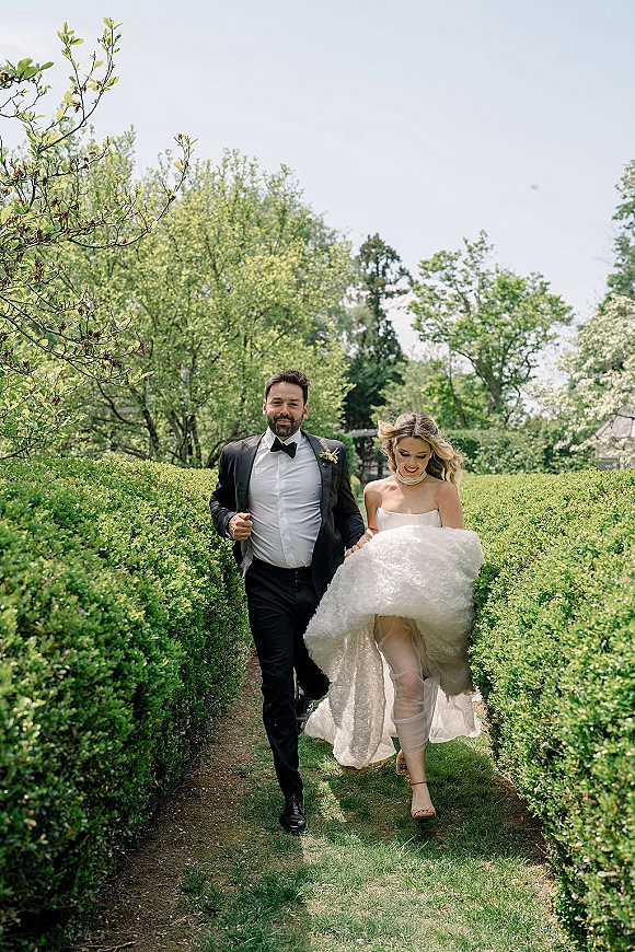 Couple portrait of bride and groom walking toward camera, bride lifting her wedding dress as he wears a tuxedo on a garden path