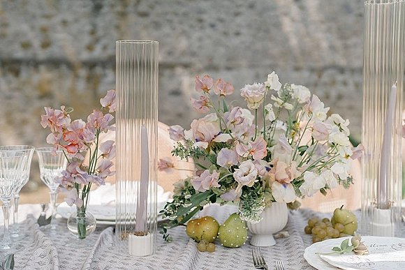 Reception tablescape with pastel wedding tablescape sweet pea centerpiece, taper candles, crystal stemware, fruit, and place card against stone wall