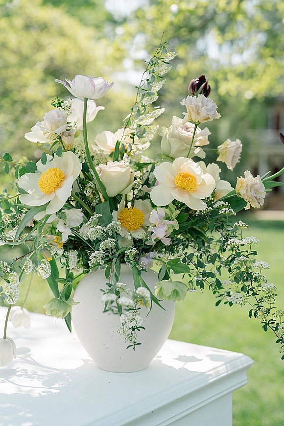 Wedding centerpiece in a white vase with airy white blooms, baby's breath and greenery on a tablecloth, garden lawn bokeh behind
