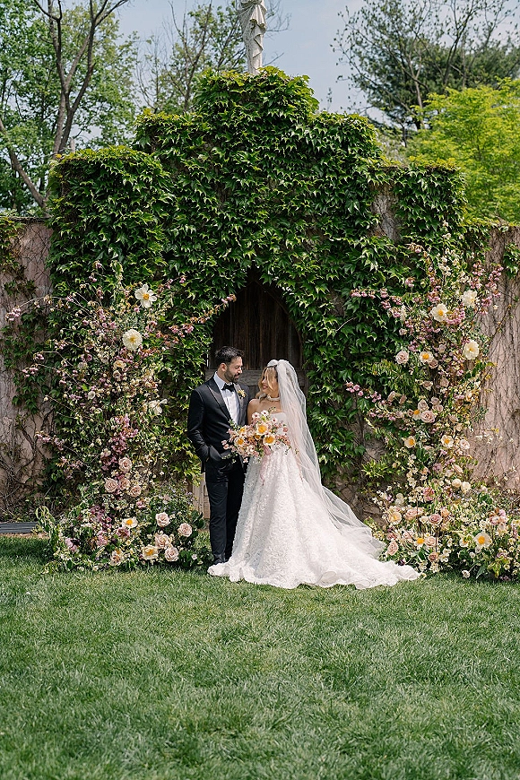 Couple portrait of bride and groom holding a colorful bouquet, gazing at each other beneath a floral arch by an ivy stone wall and door