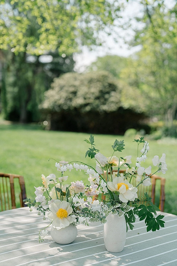 Wedding centerpiece with white flower centerpiece in ceramic vases, garden blooms and greenery on a striped tablecloth set on a lawn under trees