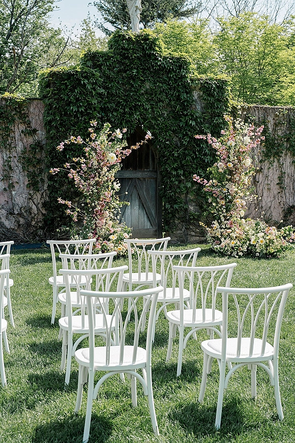 Ceremony setup with outdoor ceremony chairs in white facing a floral arch by an ivy-covered stone wall and wooden door in daylight