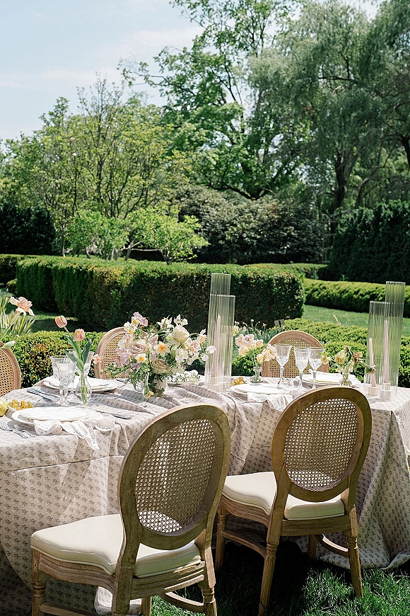 Reception tablescape with an outdoor reception table set in pastel tulips, taper candles, ribbed glass hurricanes, and cane-back chairs on a garden lawn