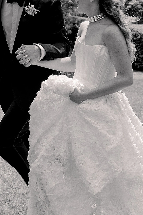 Wedding couple portrait of bride and groom holding hands, strapless floral appliqué ball gown and tuxedo on a lawn with hedges