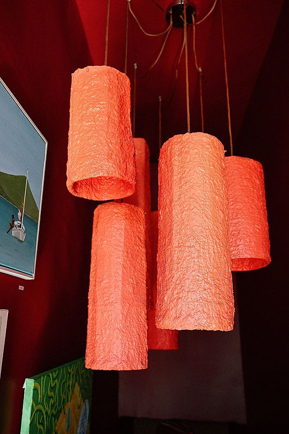 Hanging lanterns in a paper lantern chandelier cluster, cylindrical shades glowing softly from cords against a red ceiling and wall