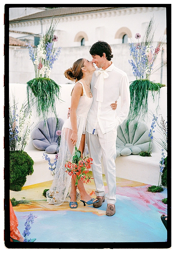 Couple portrait of bride and groom embrace nose to nose, her bouquet with orange flowers on a white stucco patio with arches