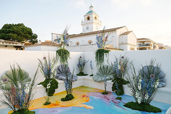 Wedding lounge decor with pastel chairs and built-in bench seating, framed by tall grass florals on moss against a white stucco wall backdrop