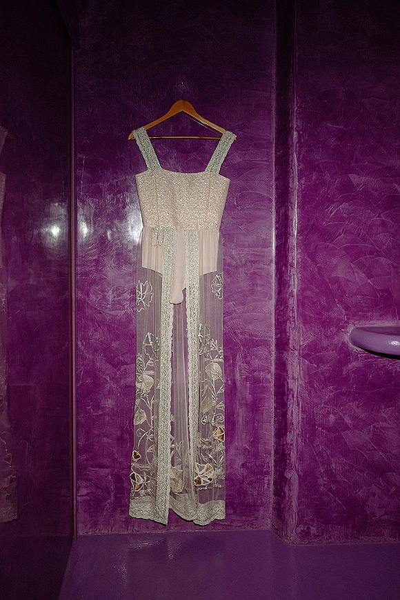 Wedding dress on hanger with glitter bodice and spaghetti straps, tulle overskirt and floral appliques against a purple textured wall alcove