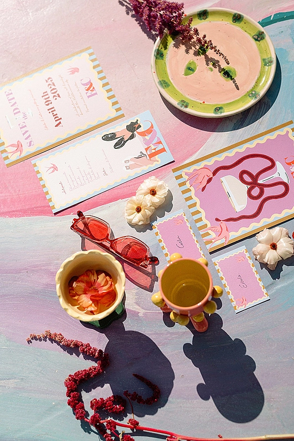 Invitation suite wedding invitation flat lay with RSVP and place cards, illustrated stationery, flowers and sunglasses on a pastel table in shadow