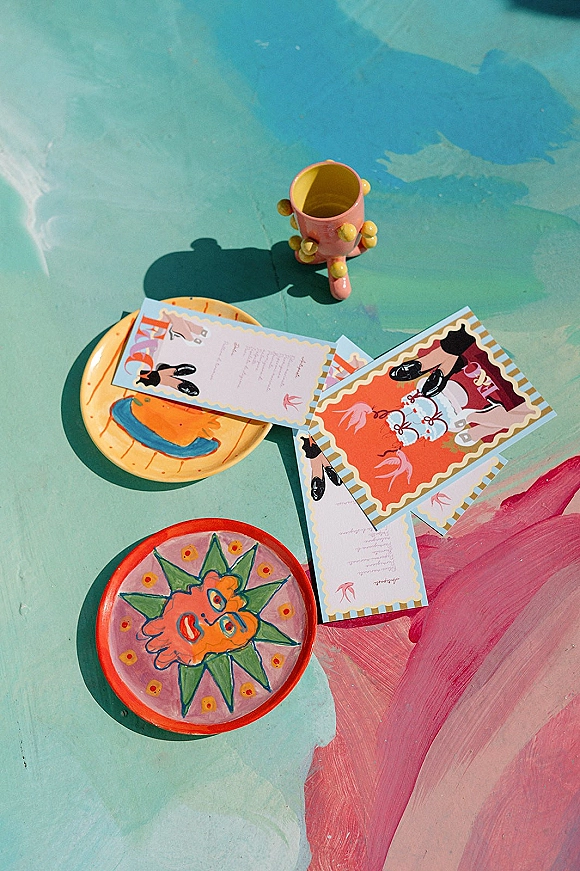 Invitation suite with colorful wedding invitations, illustrated RSVP cards, envelopes and ceramics on a painted tabletop in sunlit shadows