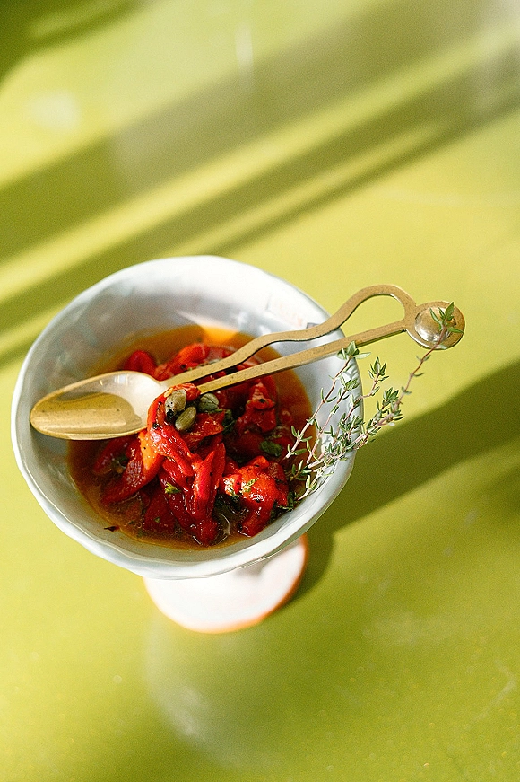 Wedding appetizer of roasted red peppers and capers in a white bowl with a herb sprig, on a green tabletop with sunlight shadows