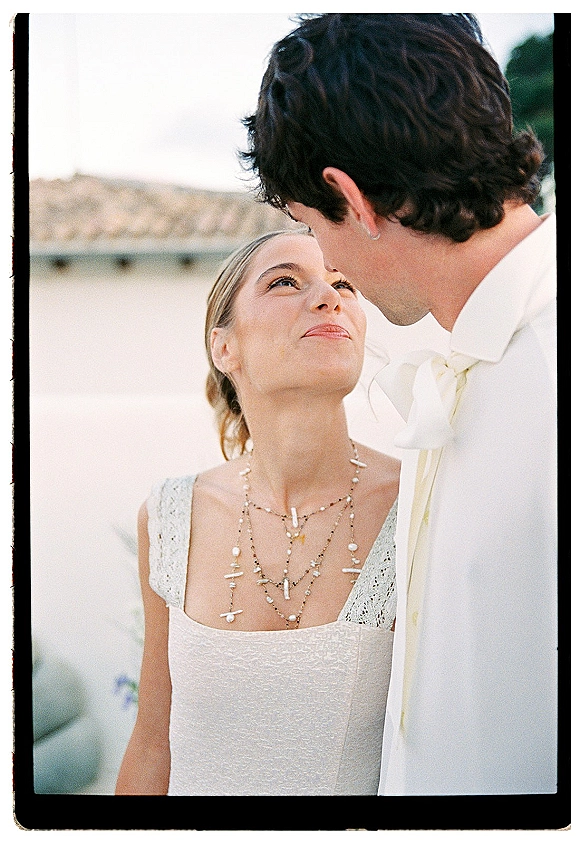 Couple portrait of bride looking up at groom, lace-strap wedding dress and layered necklaces, groom in white suit by stucco wall