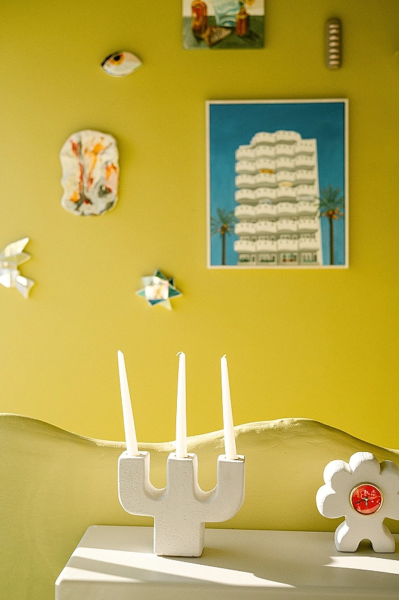 Modern candlesticks and a white ceramic candlestick holding three taper candles on a sunlit shelf ledge against a yellow wall with art prints