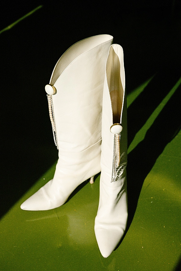 Bridal boots in white wedding boots style with pointed toes, kitten heels, and pearl tassel accents on a green floor by a dark wall