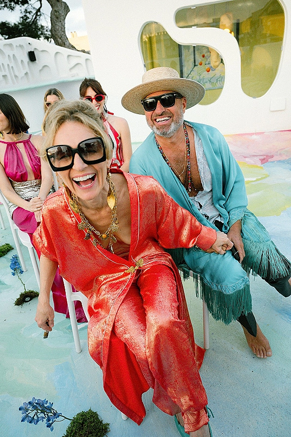 Wedding guest portrait in colorful wedding guest outfit wearing wide brim hat and sunglasses, standing on a patio by a white stucco wall